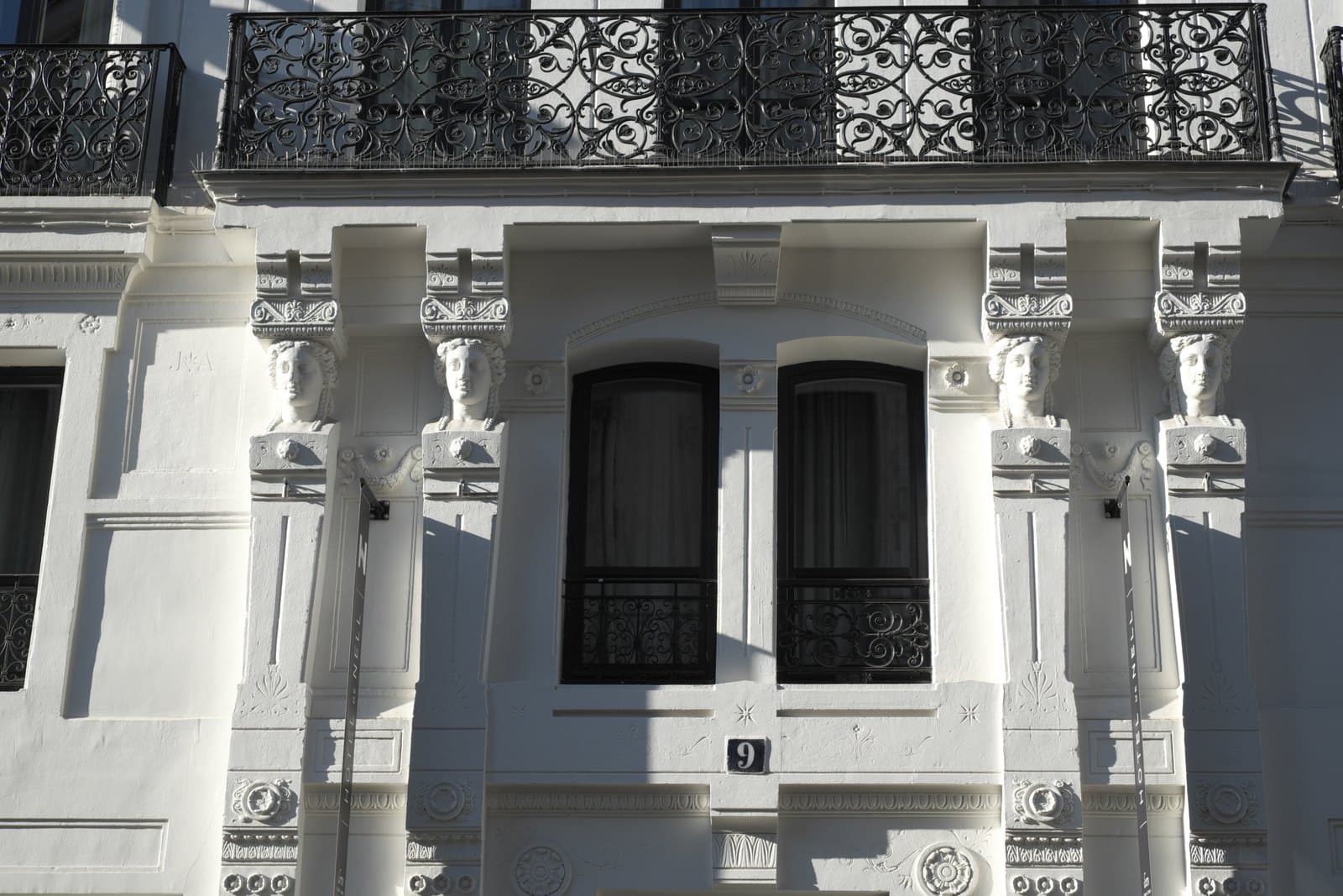 Close photograph of two pairs of carved female busts flanking twin arched windows on the white façade of 9 rue du Conservatoire, below an ornate iron balcony.