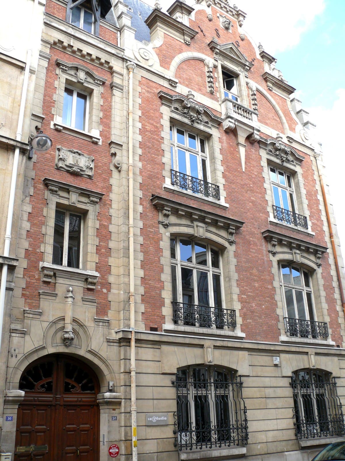 Photograph of the same brick-and-stone front in 2011, before the hotel, with a wooden carriage door between plaques numbered 30 and 28, the architect's signature carved to the left of the arch and a small sign reading le Studio beside a barred window.