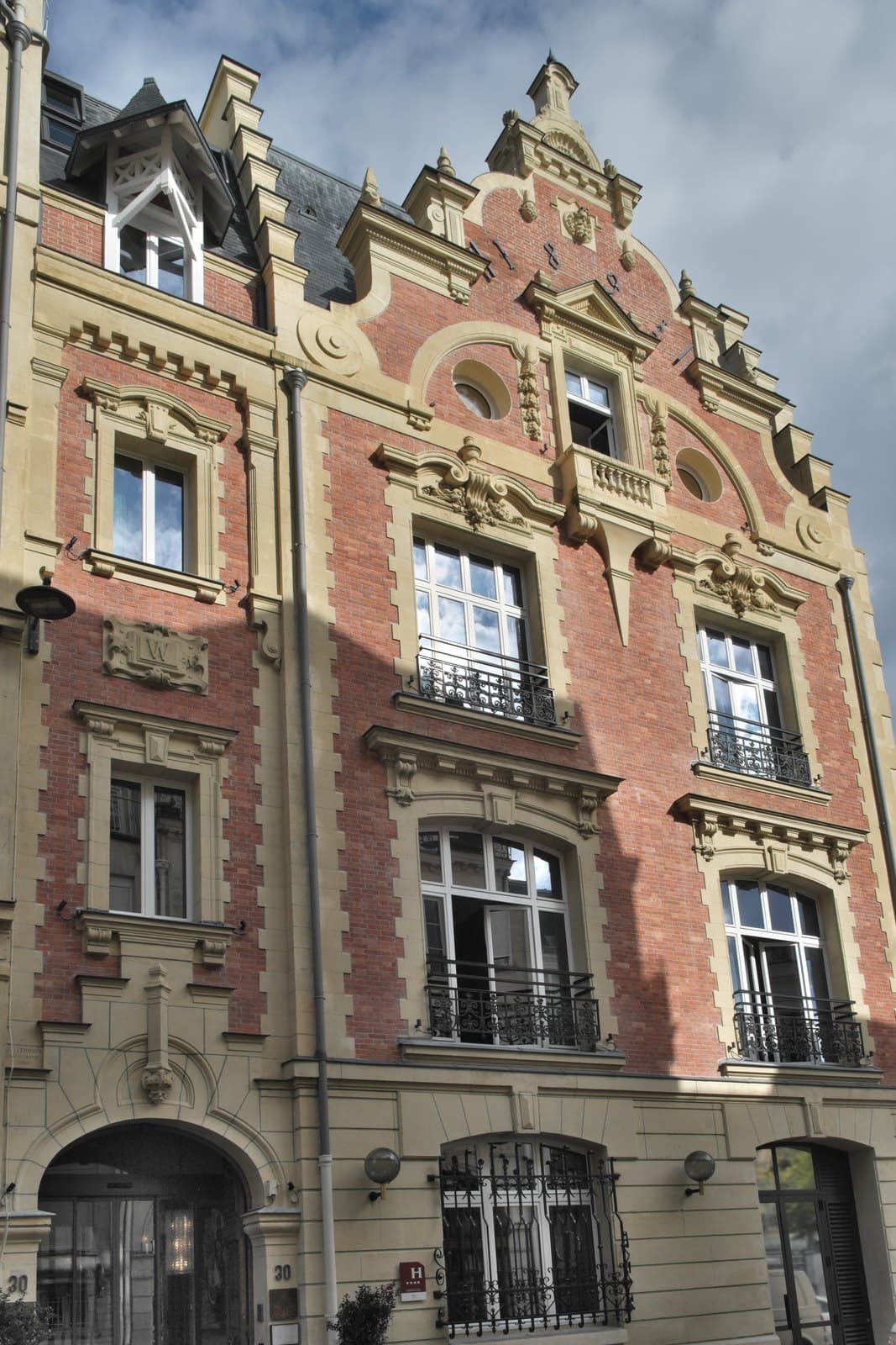 Photograph looking up the front of Le Ballu under a cloudy sky, from the arched entrance numbered 30 and a blue hotel-classification plaque to the gable, where iron anchors form the building's date between two round windows.