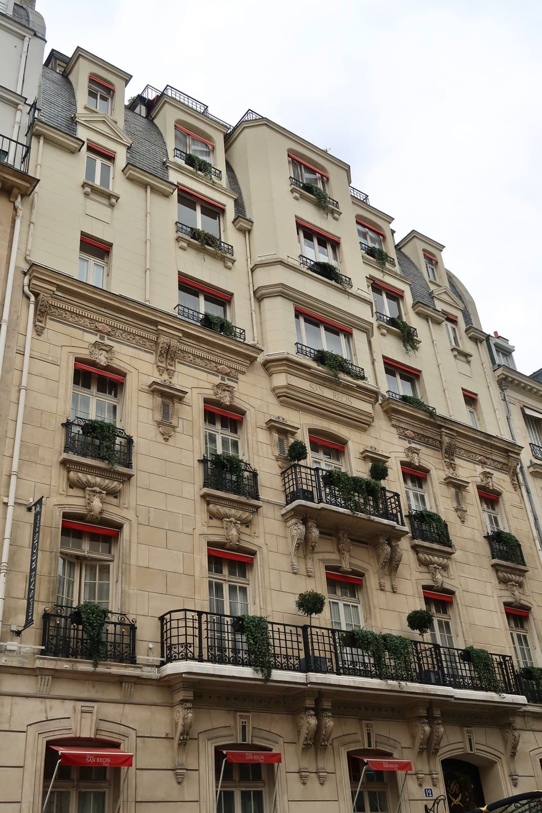 Photograph looking up the stone façade of the Hôtel San Régis on an overcast day, with planted balconies, red window awnings and red ground-floor awnings printed with the hotel's name.