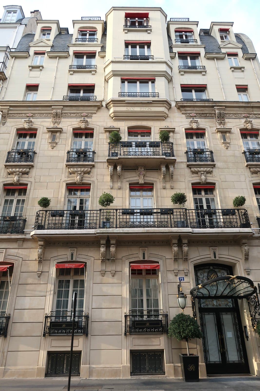 Photograph of the cut-stone front of the Hôtel San Régis: carved balconies with iron railings and small topiary trees, red-lined awnings over the windows, set-back upper storeys, and a black ironwork canopy over the entrance at street level.