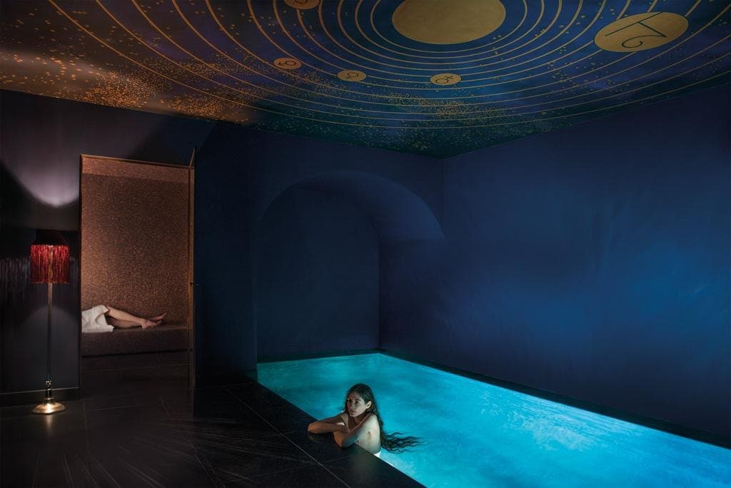 Photograph of the Salon d'Eau: a long, lit turquoise pool in a dark blue room under a ceiling painted with gold planetary rings and stars, with a woman resting her arms on the pool's edge, a fringed red lamp and a woman lying in a tiled alcove.