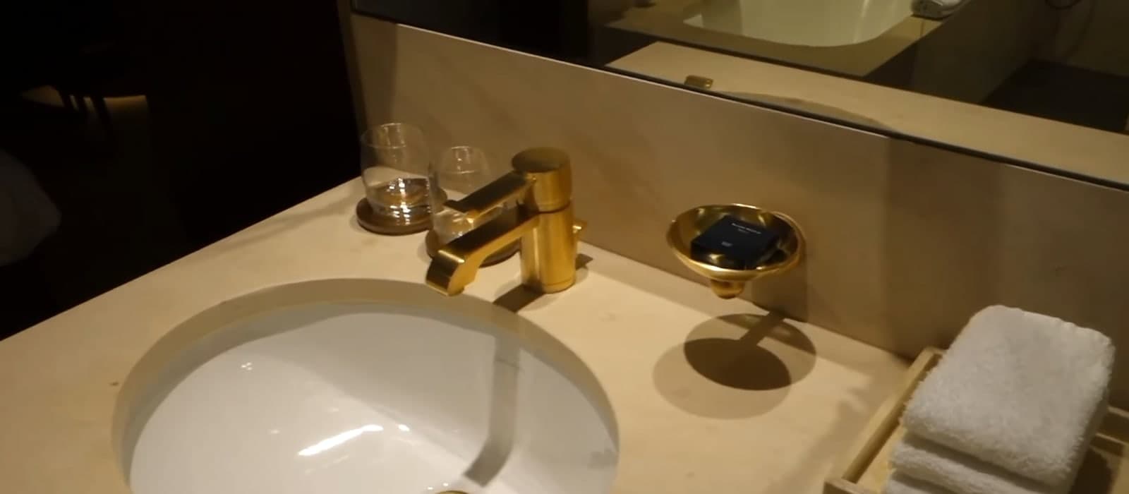 Photograph of a bathroom basin set into a pale stone counter, with a brushed-gold tap, two glasses, a gold soap dish and folded white towels below a long mirror.