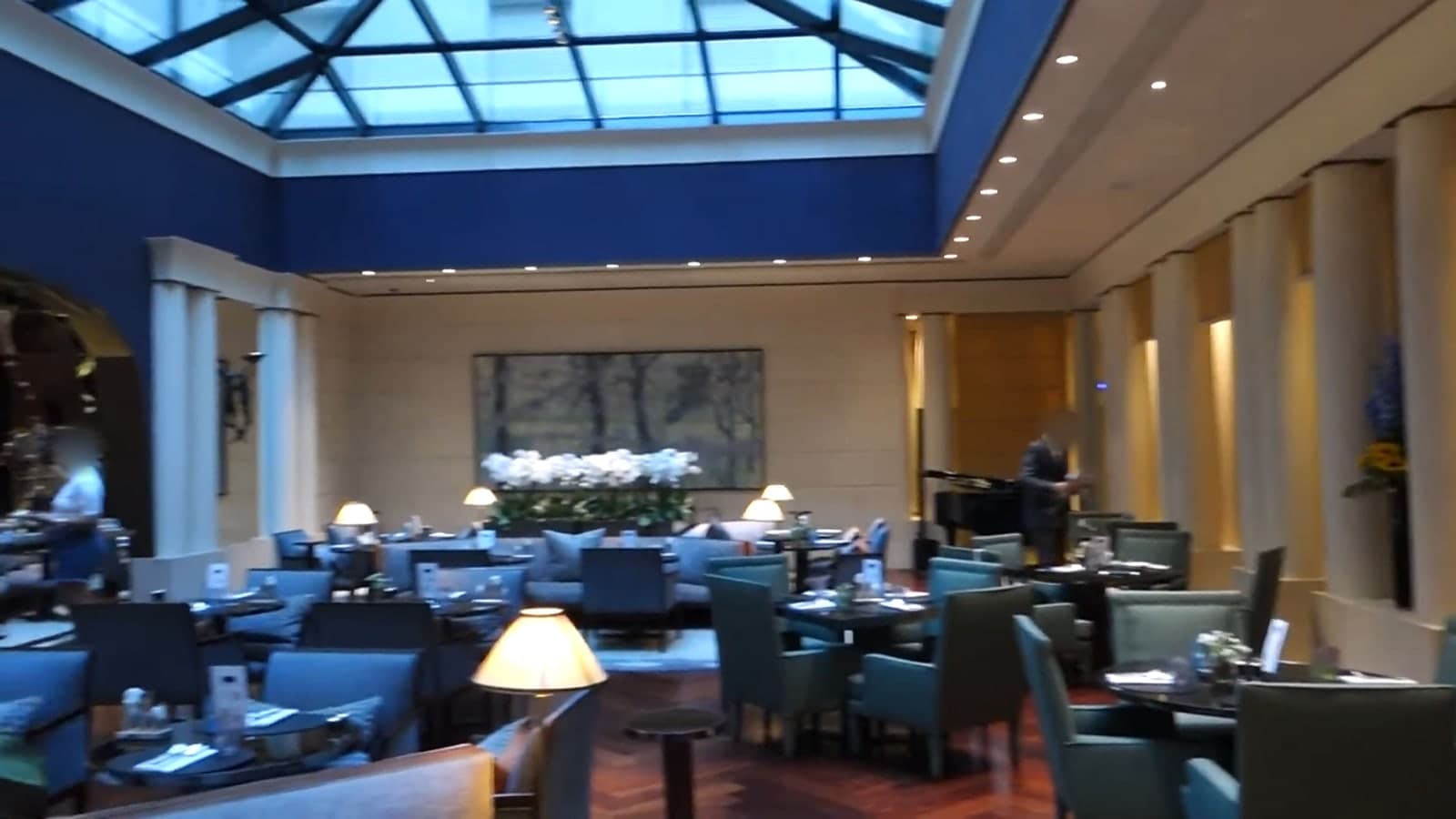 Photograph of a large dining room under a glass roof, with deep blue upper walls, cream columns, a long display of white orchids in front of a painting, and tables with blue-green armchairs.