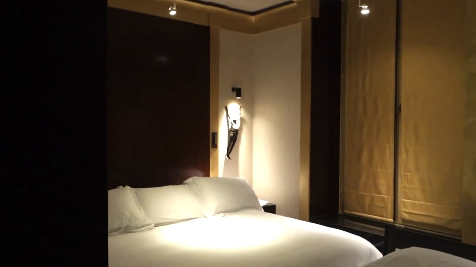 Photograph of a hotel room in low light: a double bed with white pillows, a small bronze figure lamp on the wall above it, and tall wooden panels with pale blinds over the window.