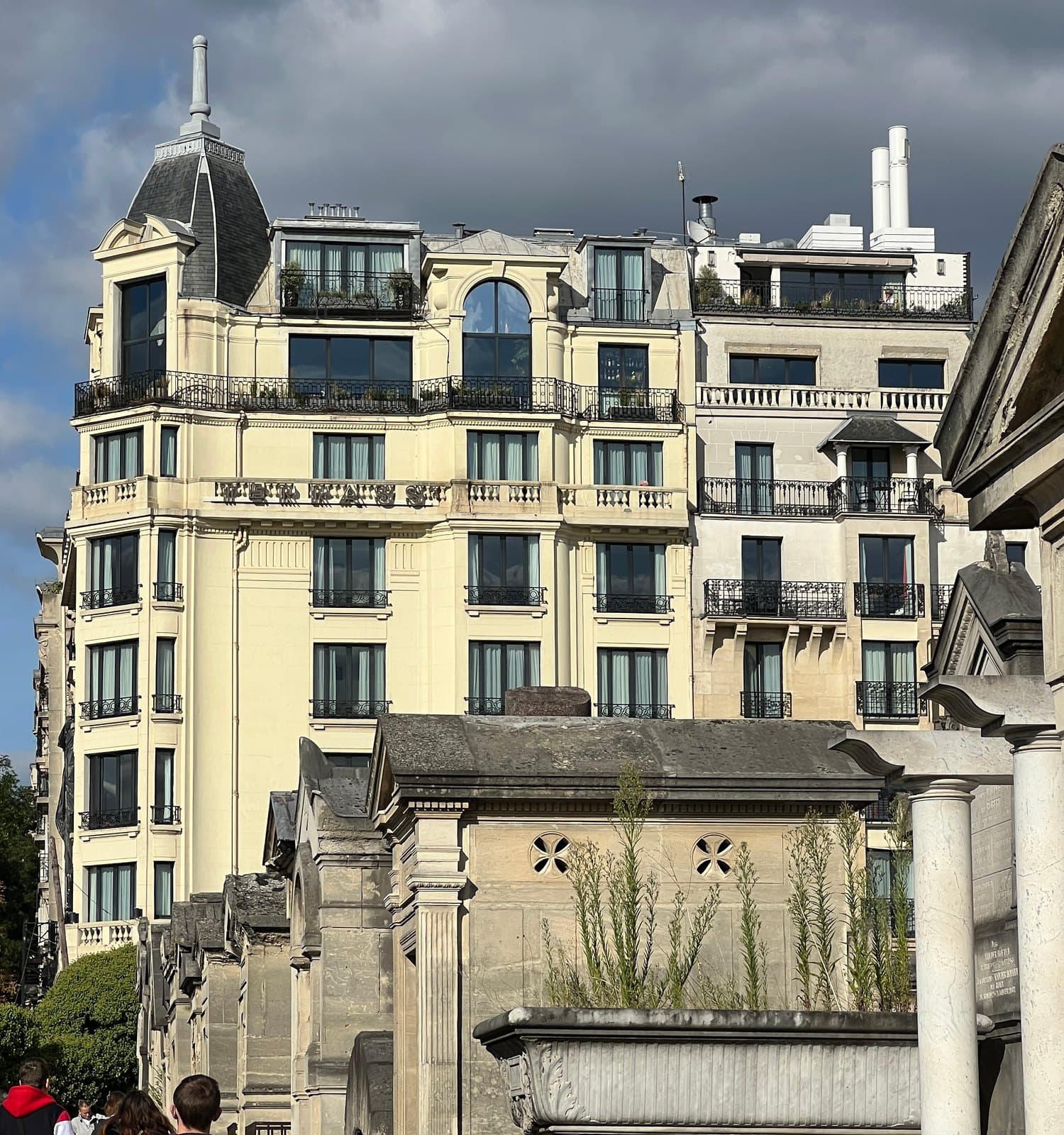 Photograph of the hotel's corner pavilion, topped by a slate dome and a finial, with cream stone walls, iron balconies and glazing on the roof, rising above the tombs of Montmartre Cemetery.