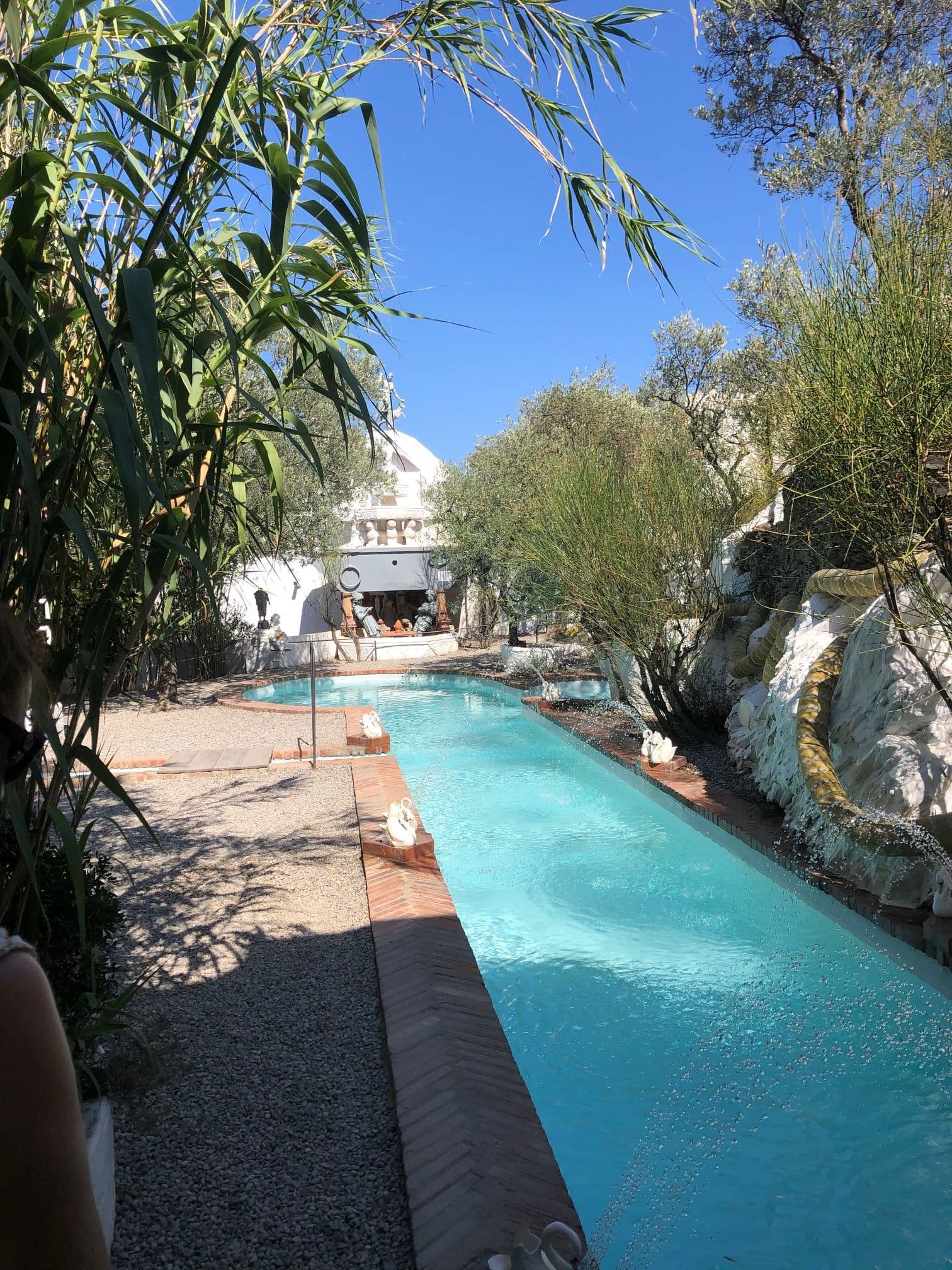 Long turquoise swimming pool in the garden of Salvador Dalí's white house at Portlligat, surrounded by reeds, olive trees, and sculptural objects