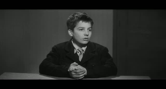 Young Jean-Pierre Léaud as Antoine Doinel seated at a table in a dark jacket and tie, hands clasped, in black and white