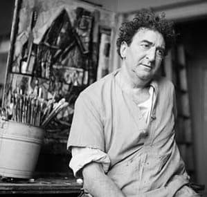 Abraham Rattner seated in his studio in a work smock, brushes in a pot beside a canvas.