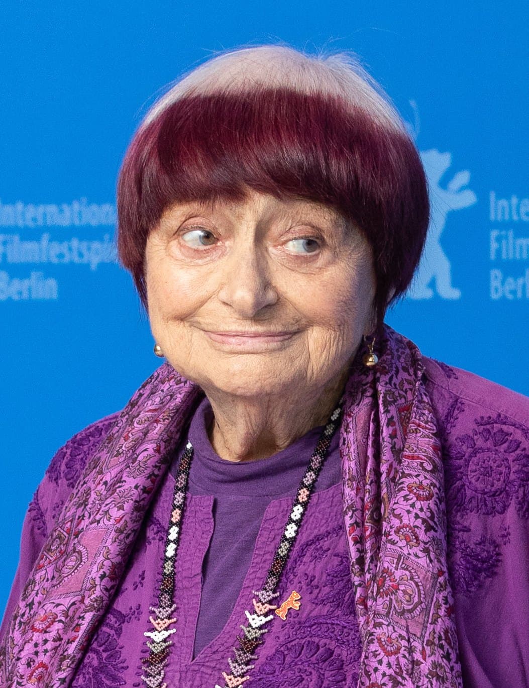 Agnès Varda in a purple jacket and scarf at the Berlinale, her bowl-cut hair white at the crown and red at the ends.