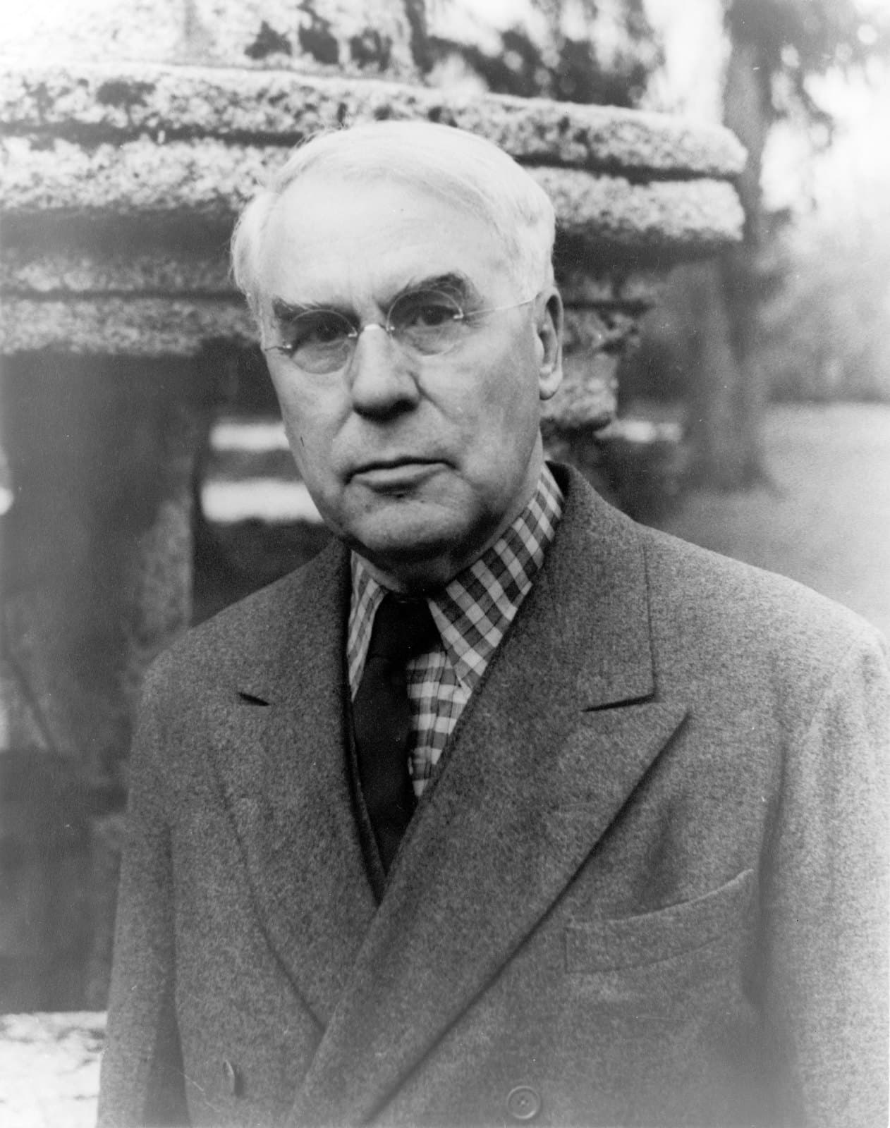 Albert C. Barnes in rimless glasses, checked shirt, and overcoat, photographed outdoors before a stone wall.