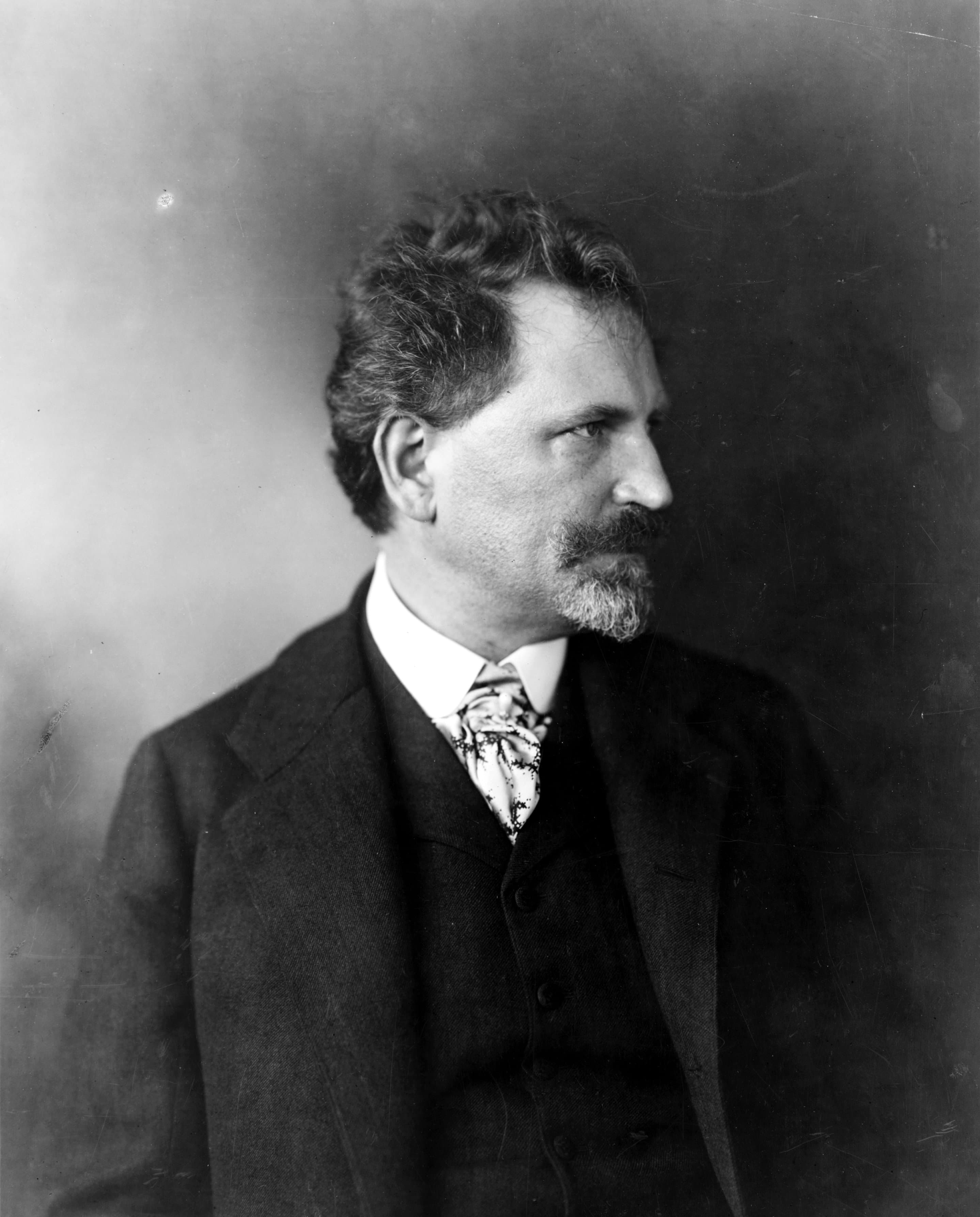 Alphonse Mucha in middle age, bearded, seated with arms crossed and looking directly at the camera