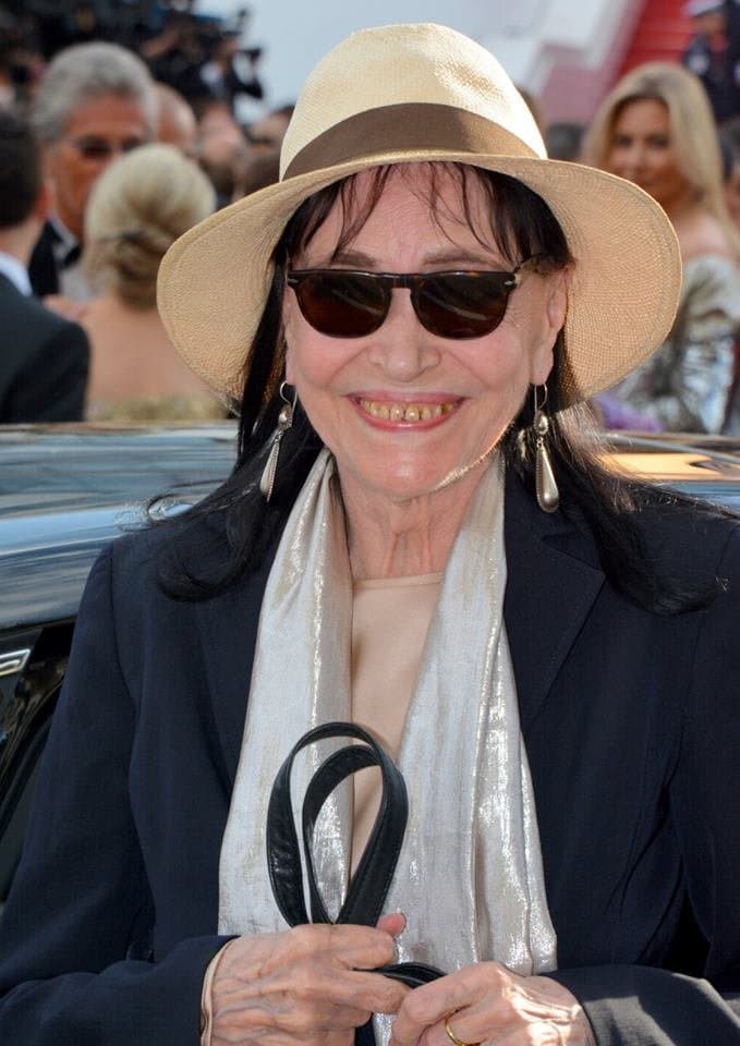 Anna Karina in a white brimmed hat and sunglasses, smiling, a silver scarf at her neck, at Cannes in 2018
