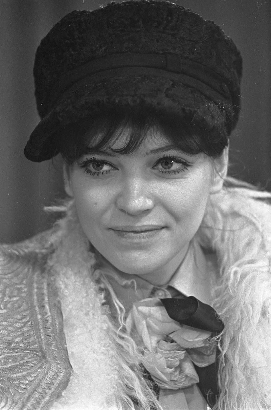 Anna Karina in a dark fur hat and patterned coat at a press conference, looking slightly off-camera, in black and white