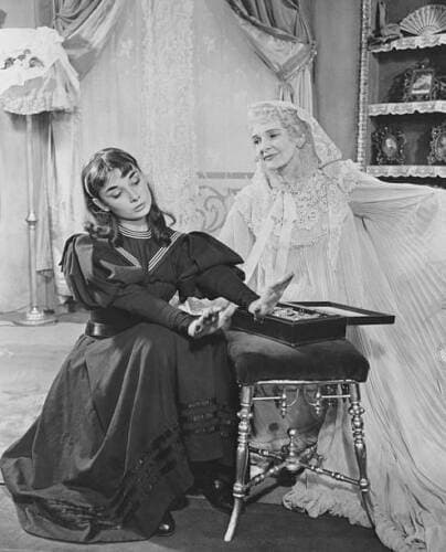 Audrey Hepburn, seated in a dark high-collared dress, receives a jewel case from Cathleen Nesbitt, who stands in an elaborate lace gown and veil, on the set of the 1951 Broadway play Gigi