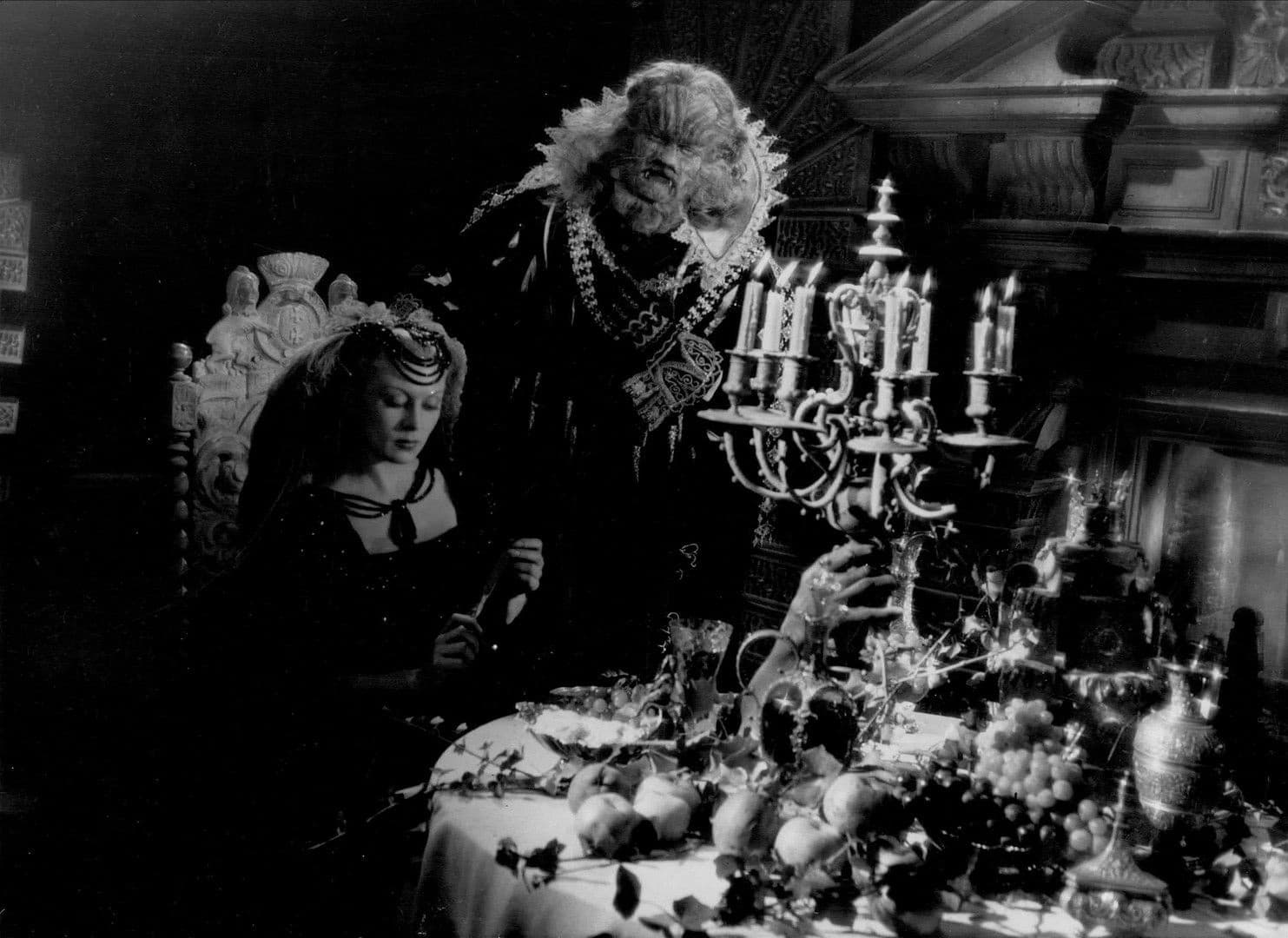 Josette Day as Belle seated at a laden banquet table while Jean Marais as the Beast, in ruff and jewels, leans over her shoulder