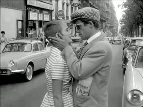 Jean-Paul Belmondo in a checked cap and sunglasses holding Jean Seberg's face in both hands on a Paris street, cars behind