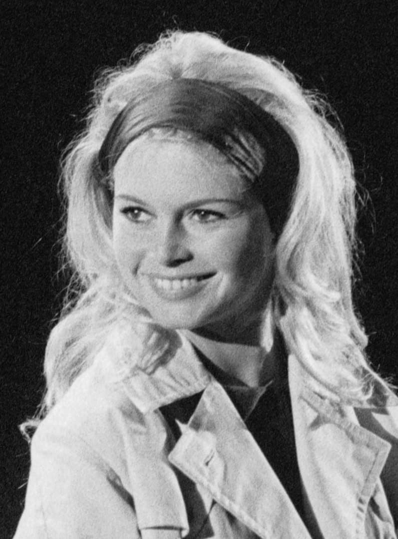 Brigitte Bardot smiling in a headband and trench coat against a dark background.