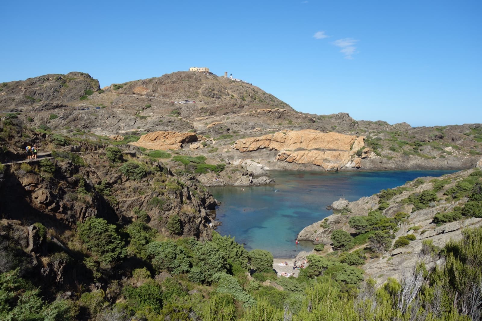 The rocky coast of Cap de Creus in Catalonia: an ochre-yellow rock outcrop above a calm turquoise cove, the cape's lighthouse on the headland beyond.