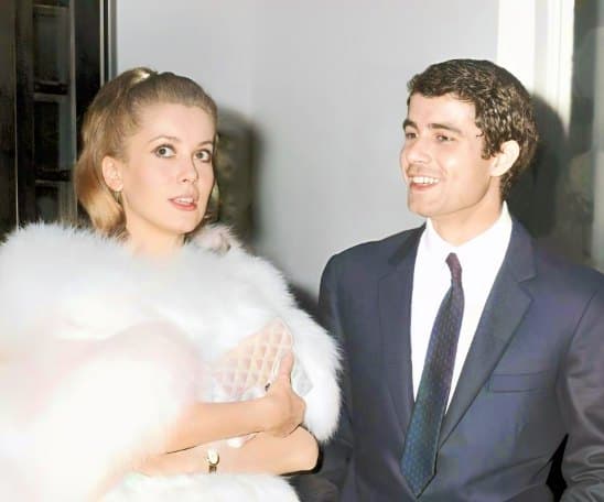Colorized press photograph of Catherine Deneuve in a white fur wrap holding a pale quilted clutch, glancing upward, beside Nino Castelnuovo grinning in a dark suit and tie