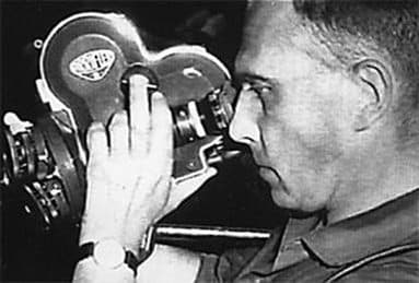 Chris Marker in profile behind a 16mm film camera, one hand on the lens turret.