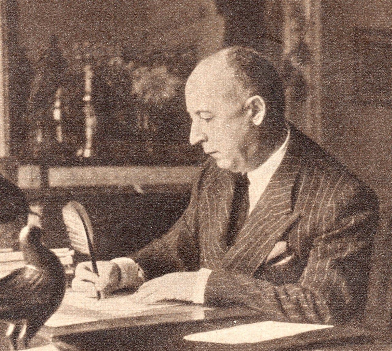 Christian Dior in 1954, seated at a desk in a dark suit with papers and a telephone in front of him.