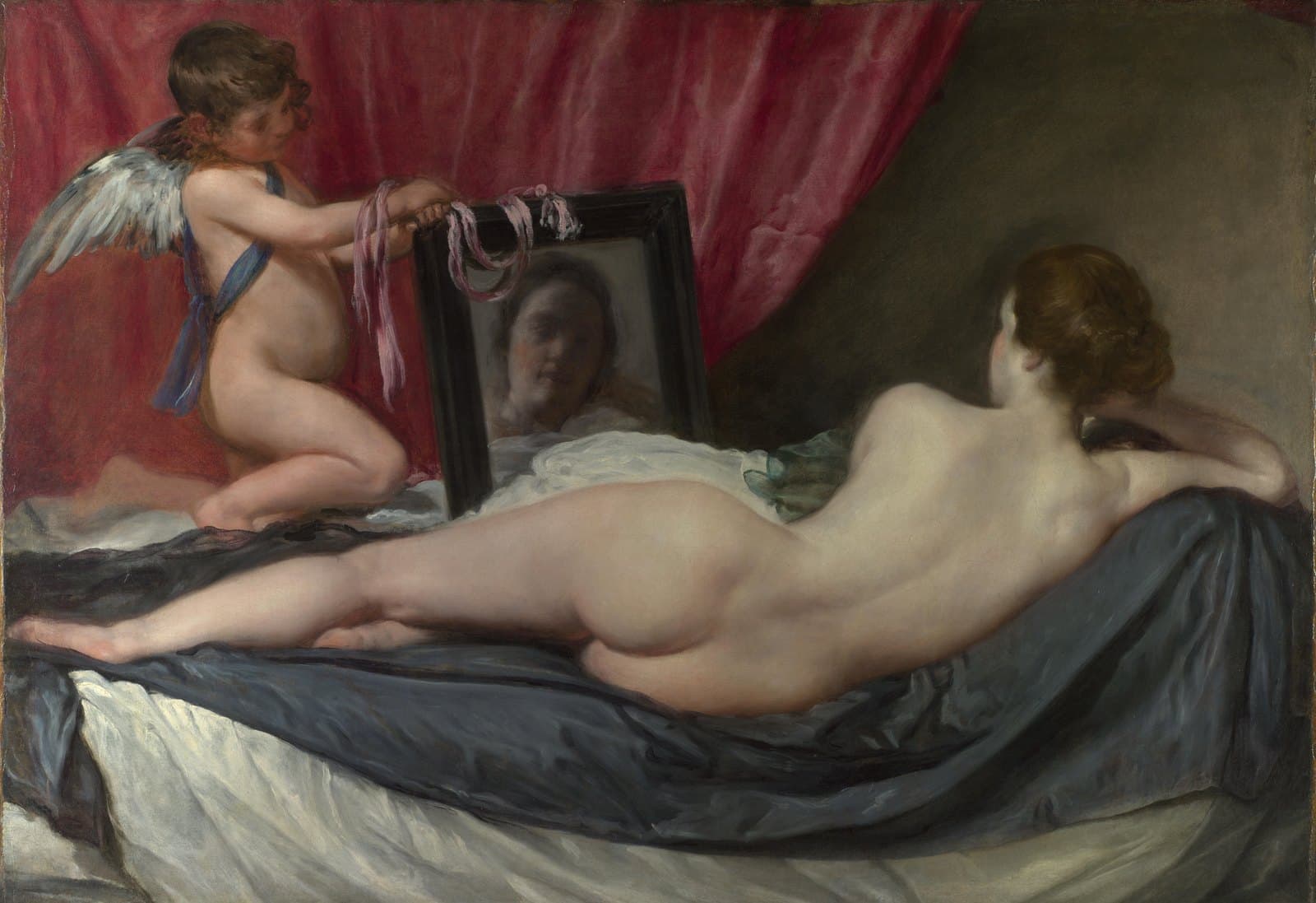A nude woman lies on her side on a bed with her back to the viewer, propped on her right elbow, her dark hair pinned up. A winged boy kneels at the foot of the bed holding up a black-framed mirror with pink ribbons draped over it, and her face appears blurred in the glass. Gray silk sheets are rumpled beneath her and a deep red curtain hangs across the upper left.