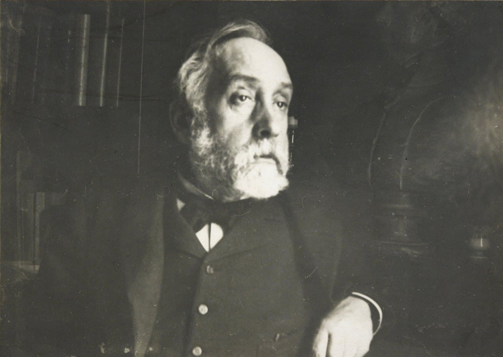 Edgar Degas, white-bearded in jacket and bow tie, lit from one side in a dim interior.