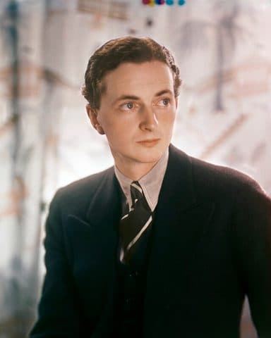 Colour studio portrait of Edward James at twenty-six, in a dark suit and striped tie against a patterned backdrop.