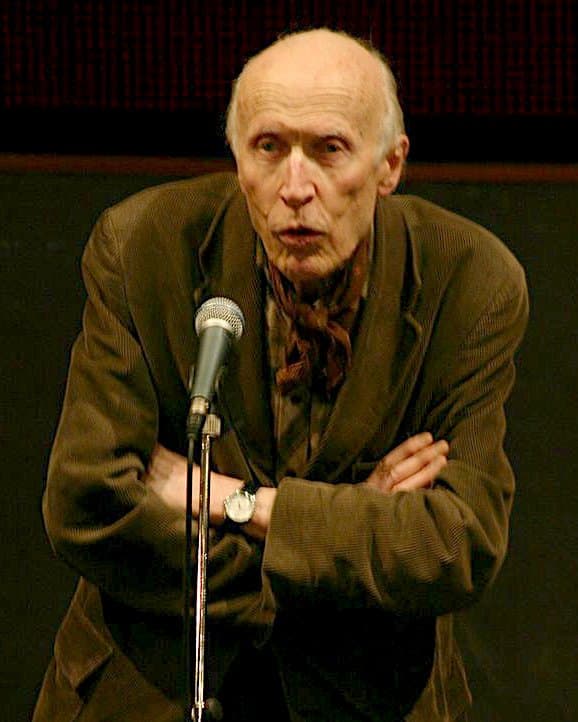 Éric Rohmer speaking at a microphone with arms crossed, in a corduroy jacket and neck scarf.