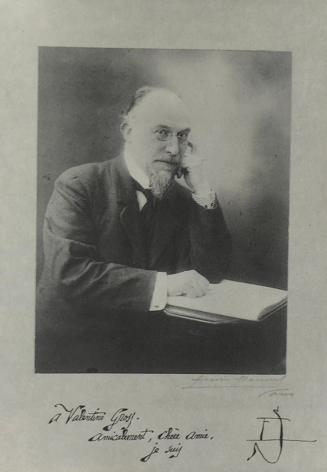 Erik Satie seated at a table with an open score, chin resting on one hand, wearing pince-nez and a dark suit; a handwritten dedication runs below the image