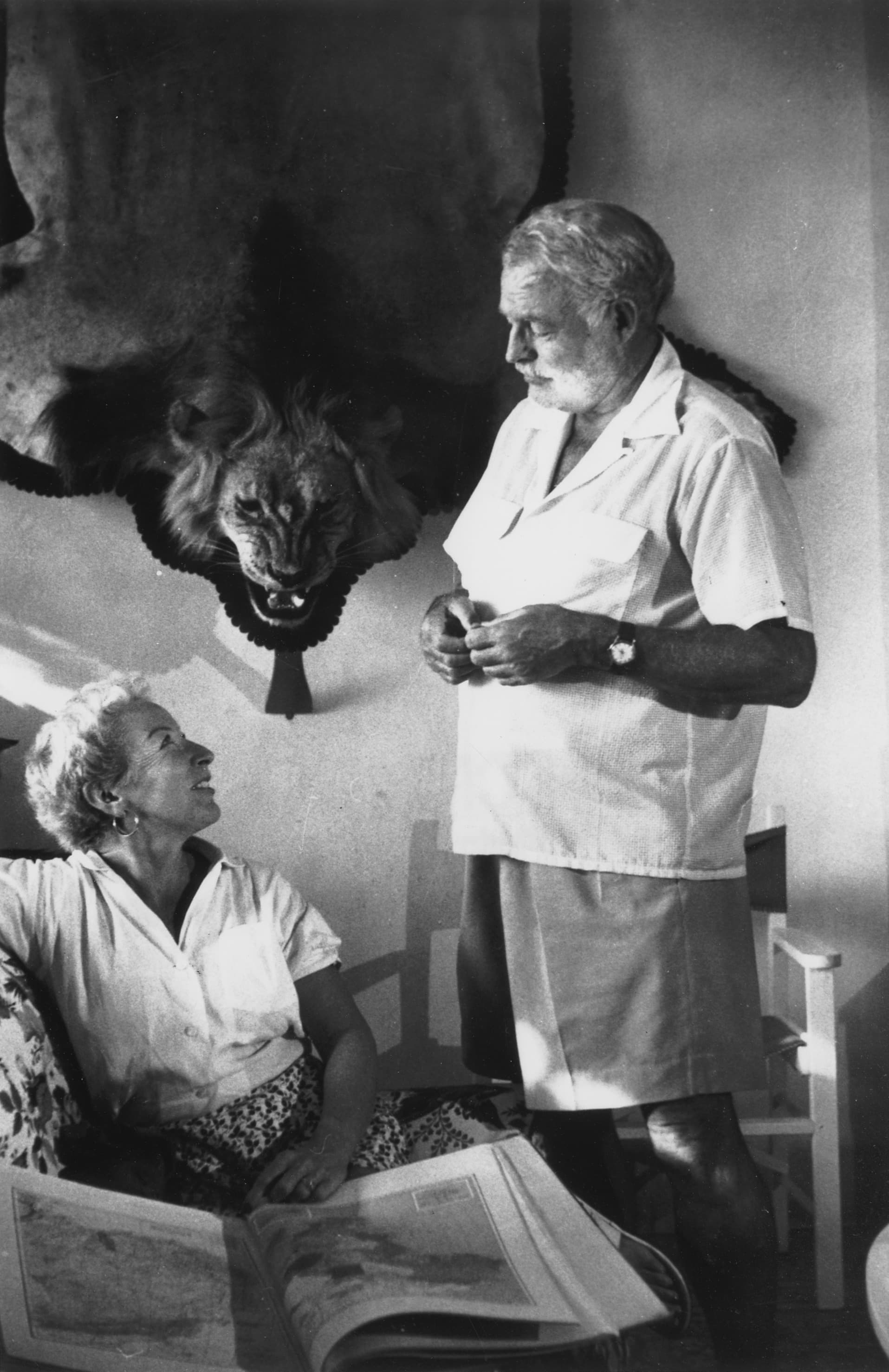 Mary Welsh Hemingway seated and Ernest Hemingway standing beneath a mounted lion's head at the Finca Vigía, Cuba