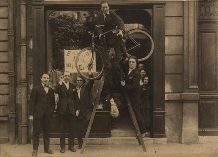 Seven members of the Paris Dada group posing at a gallery entrance, one on top of a ladder holding a bicycle, one hanging upside down, André Breton at the right