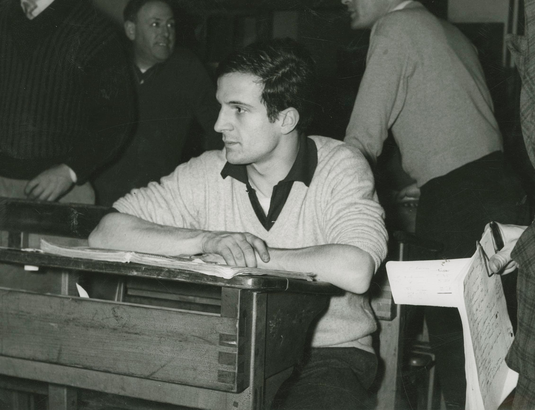 François Truffaut in a pale sweater seated at an old wooden school desk with papers, people standing behind him