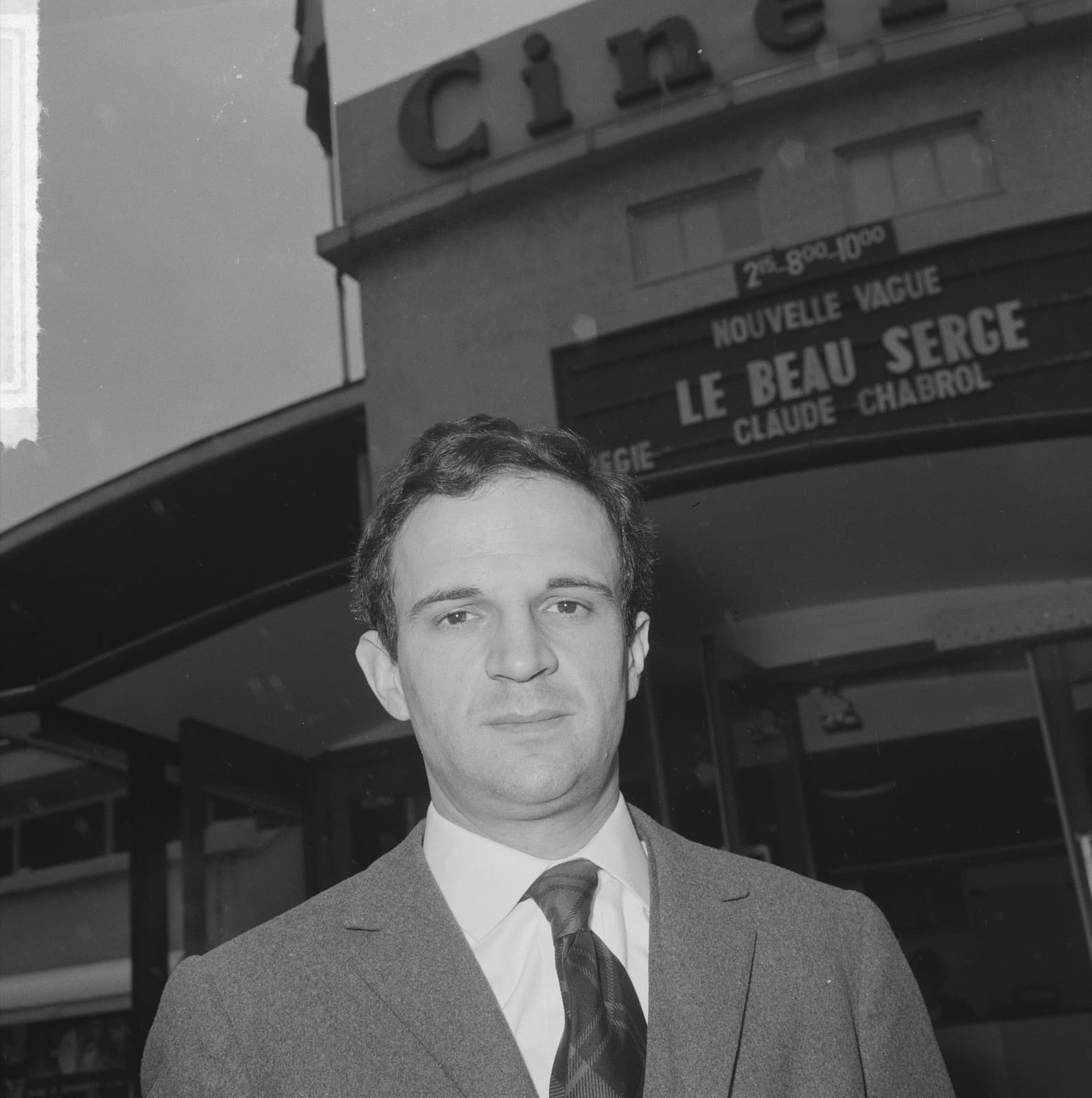François Truffaut in a grey suit and tie in front of a cinema, the marquee behind him reading Nouvelle Vague, Le Beau Serge, Claude Chabrol