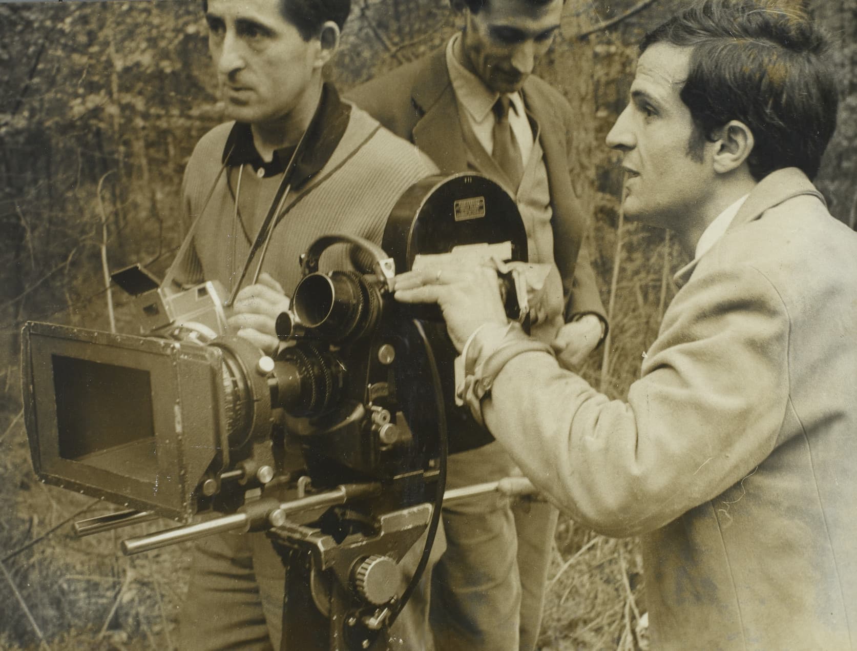 François Truffaut adjusting a 35 mm film camera outdoors, two crew members beside him among bare trees