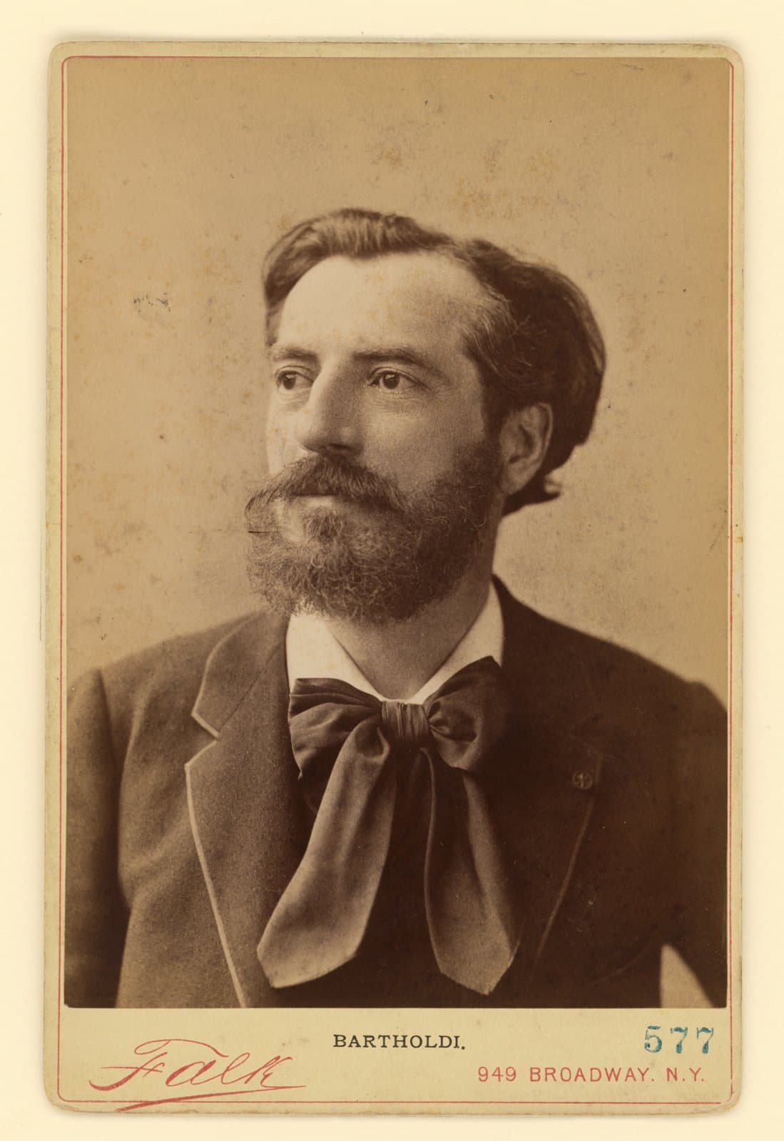 Studio portrait of Frédéric Auguste Bartholdi in 1876, a bearded man of forty-two in a tweed jacket and a wide dark bow tie, turned three-quarters to his right; the mount is printed BARTHOLDI and Falk, 949 Broadway, N.Y.