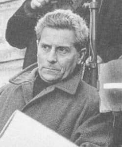Georges Franju, silver-haired in a dark coat, glancing aside on a film set.