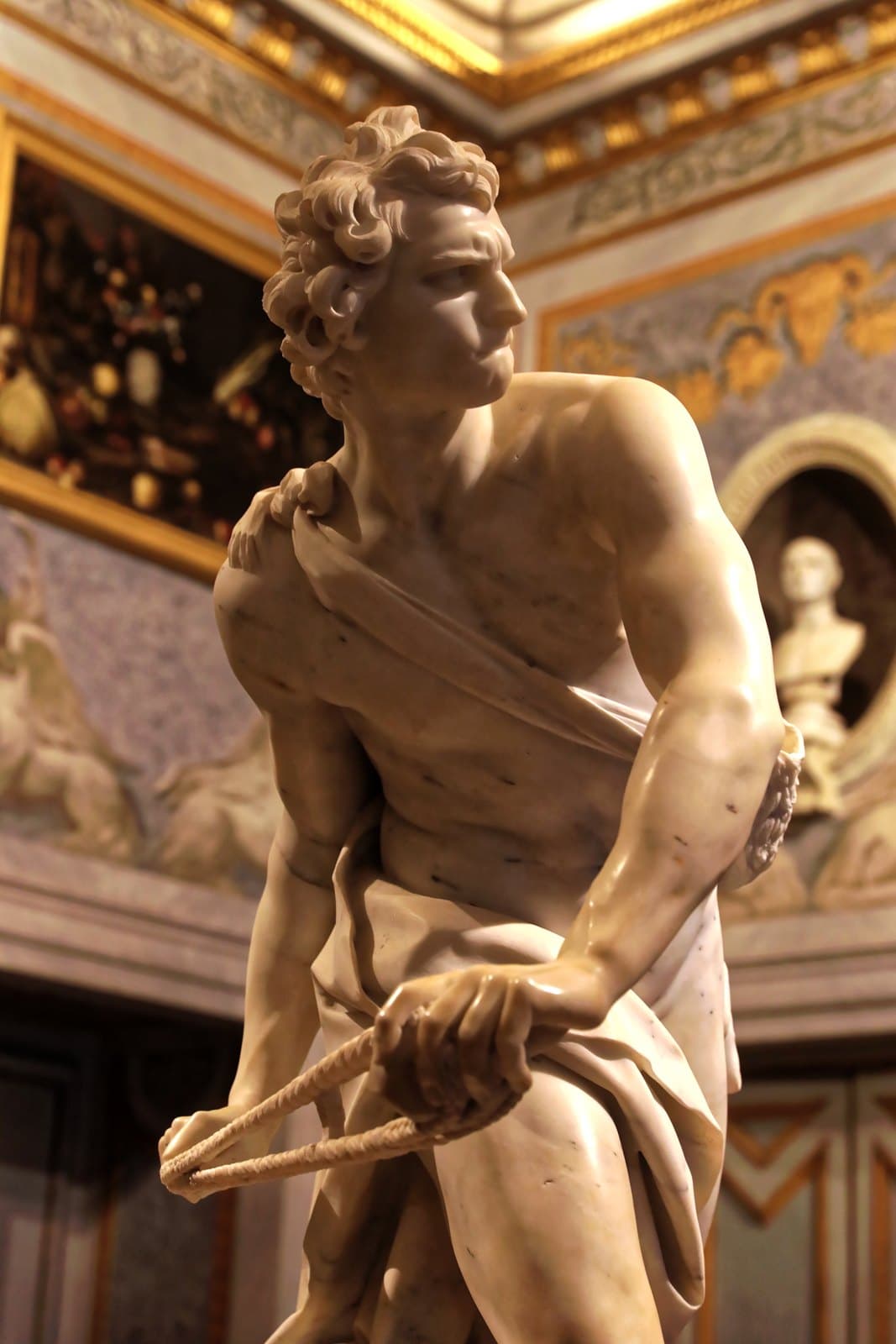 Marble figure of a young man seen from below, twisting hard to his right with a leather sling drawn across his body in both hands, his lower lip pulled in under his teeth and his eyes fixed on something outside the statue, a gilded picture frame and a marble bust in a niche behind him