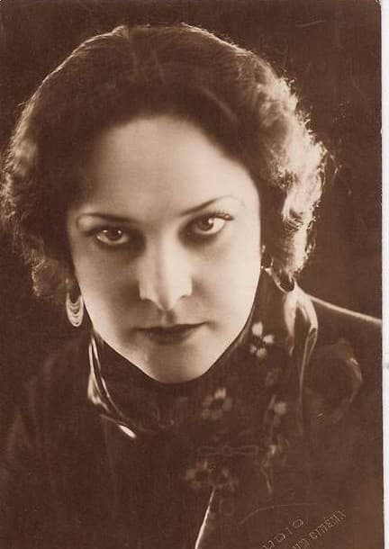 Studio portrait of Gina Manès looking directly into the camera, dark eyes heavily lit, wearing an embroidered collar