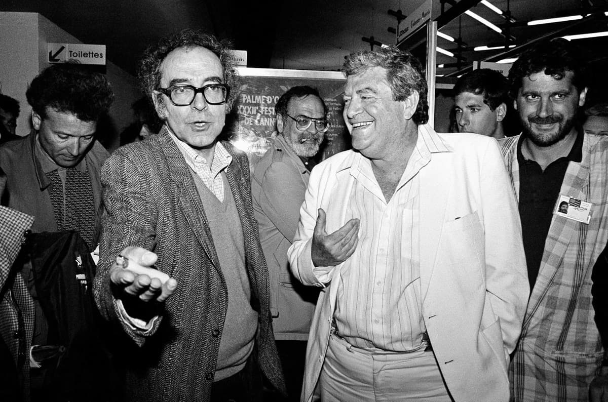 Godard gesturing in a crowded festival hall beside a laughing man in a white suit