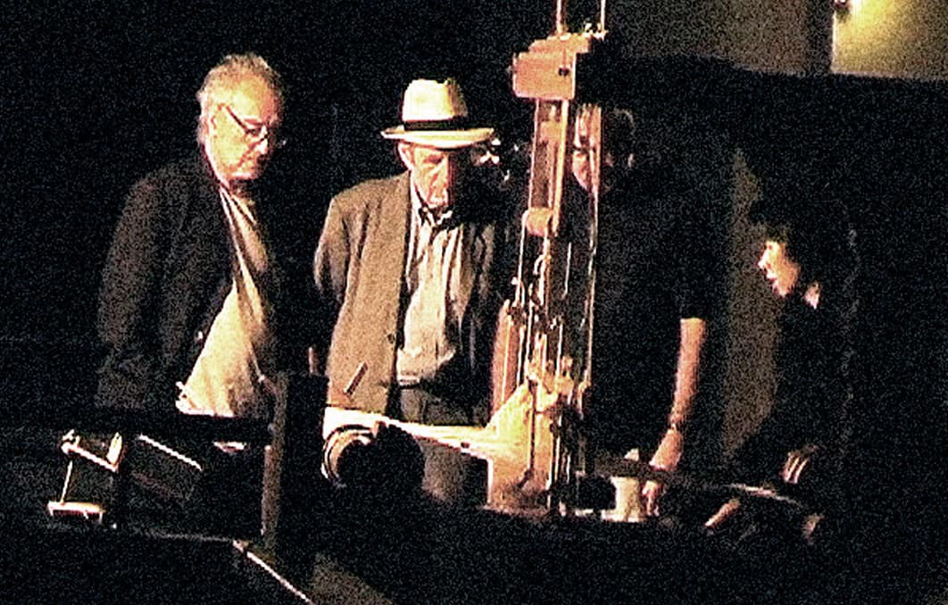 Four men in low light around a wooden camera rig in a studio; Godard wears a pale hat