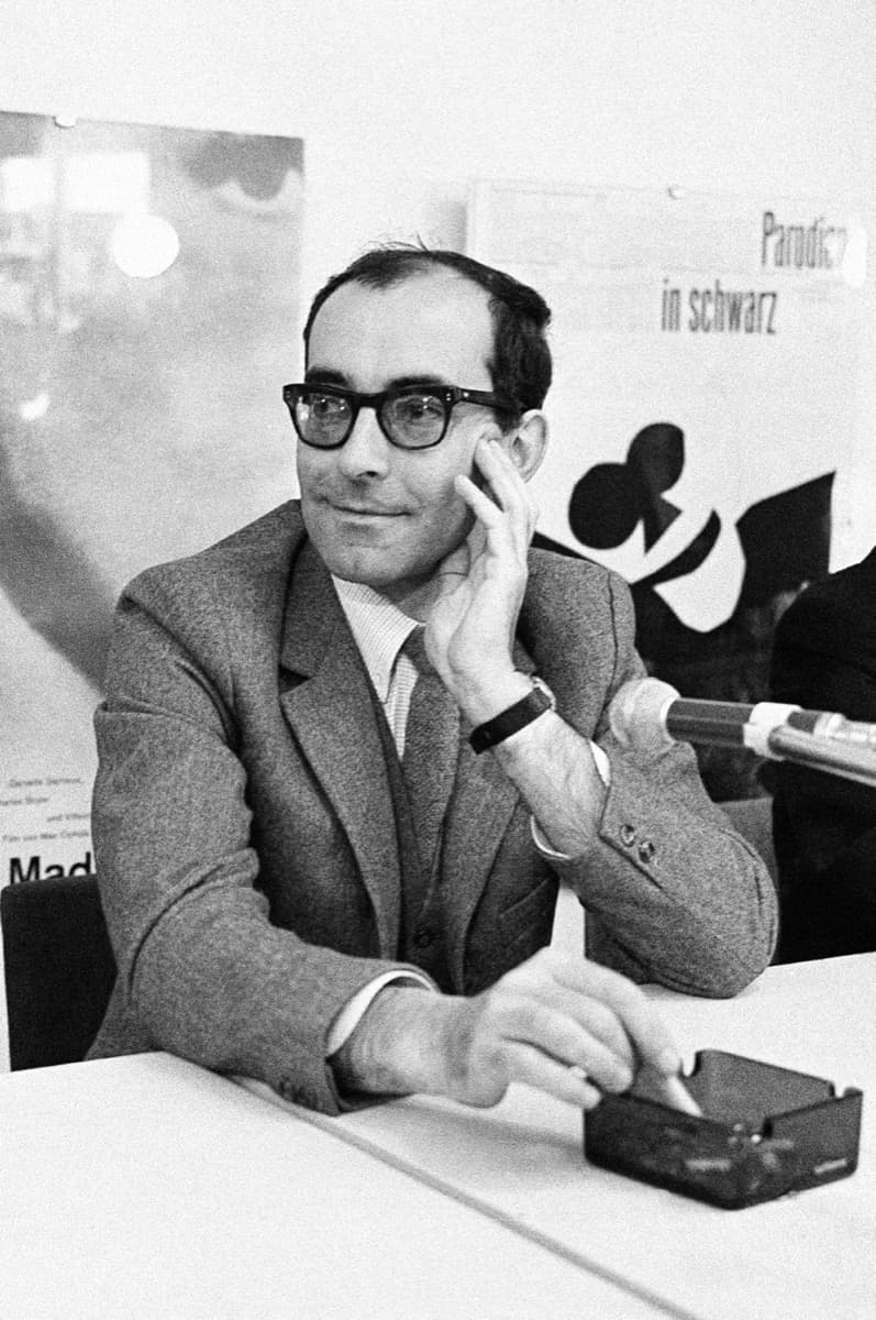 Jean-Luc Godard seated at a press table, chin resting on his hand, German-language posters behind him