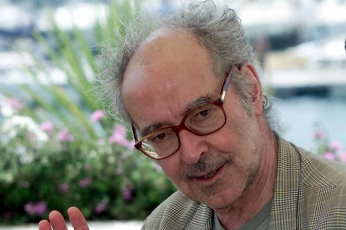 Grey-haired Godard in a plaid jacket gesturing at an outdoor photocall, harbor and flowers out of focus behind