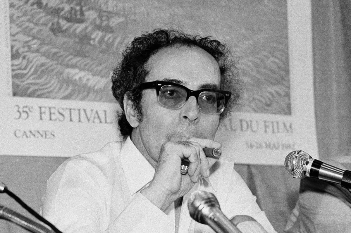 Godard in dark glasses holding a cigar at a press conference, a 35th Festival de Cannes poster behind him