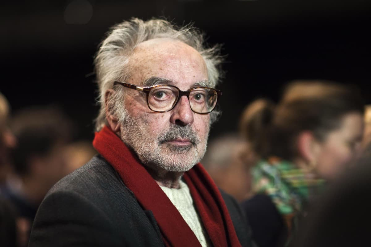 White-haired Godard in tortoiseshell glasses and a red scarf, photographed at an indoor event