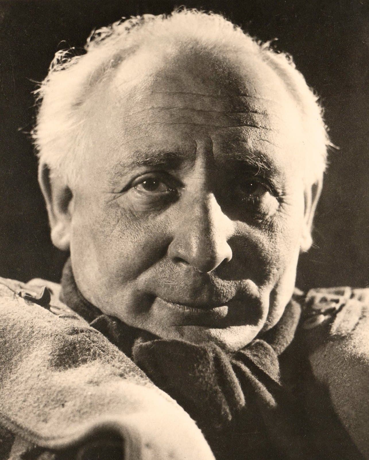 Grégoire Michonze in close-up, white-haired in a heavy coat, lit from the side against a dark background.