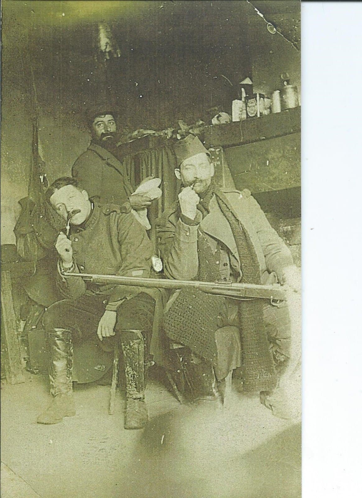 Three French soldiers seated in a billet, two smoking pipes, a rifle laid across their knees; a third man stands behind them