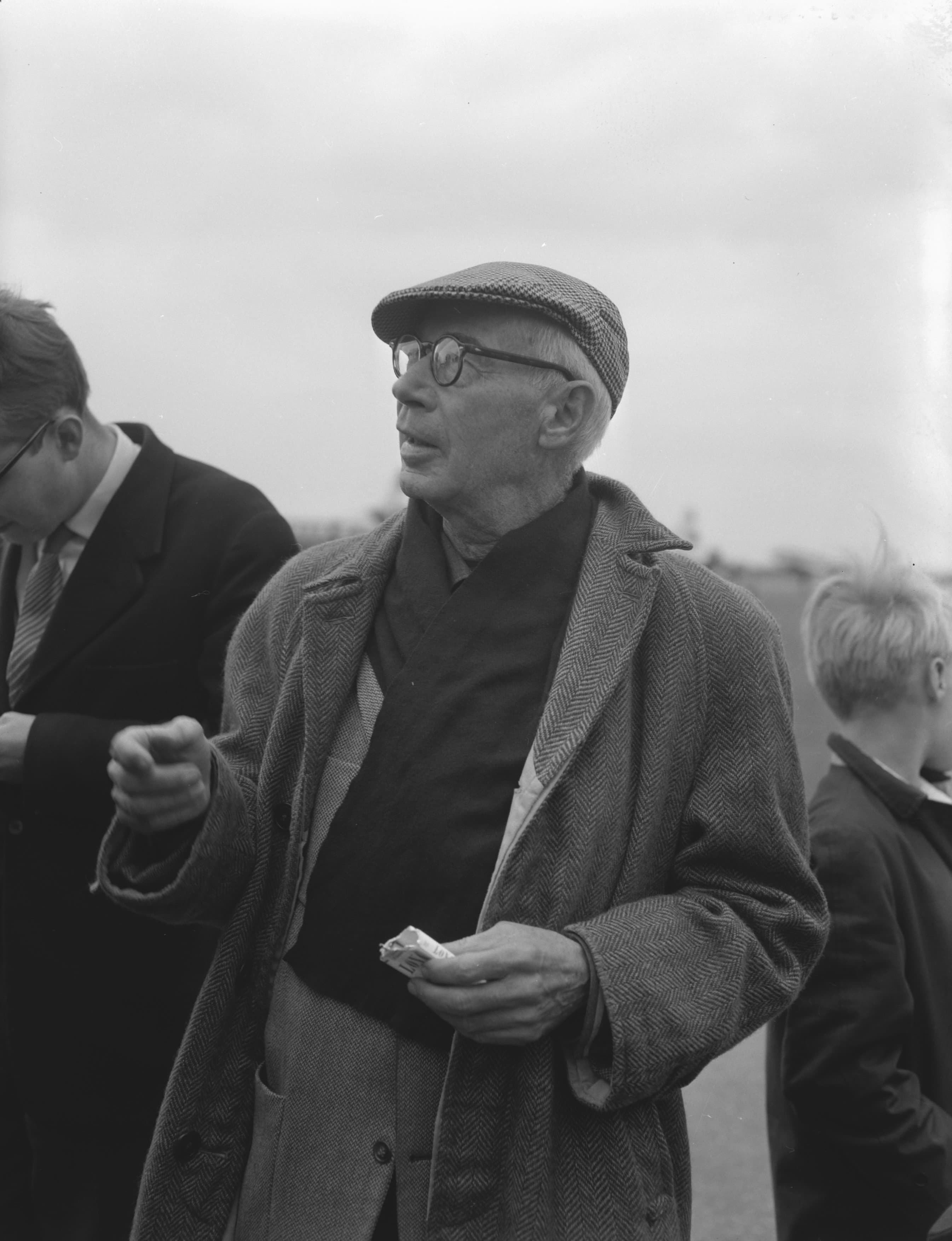 Henry Miller at sixty-seven in a flat cap and tweed overcoat, photographed among other travelers outdoors at Schiphol airport.