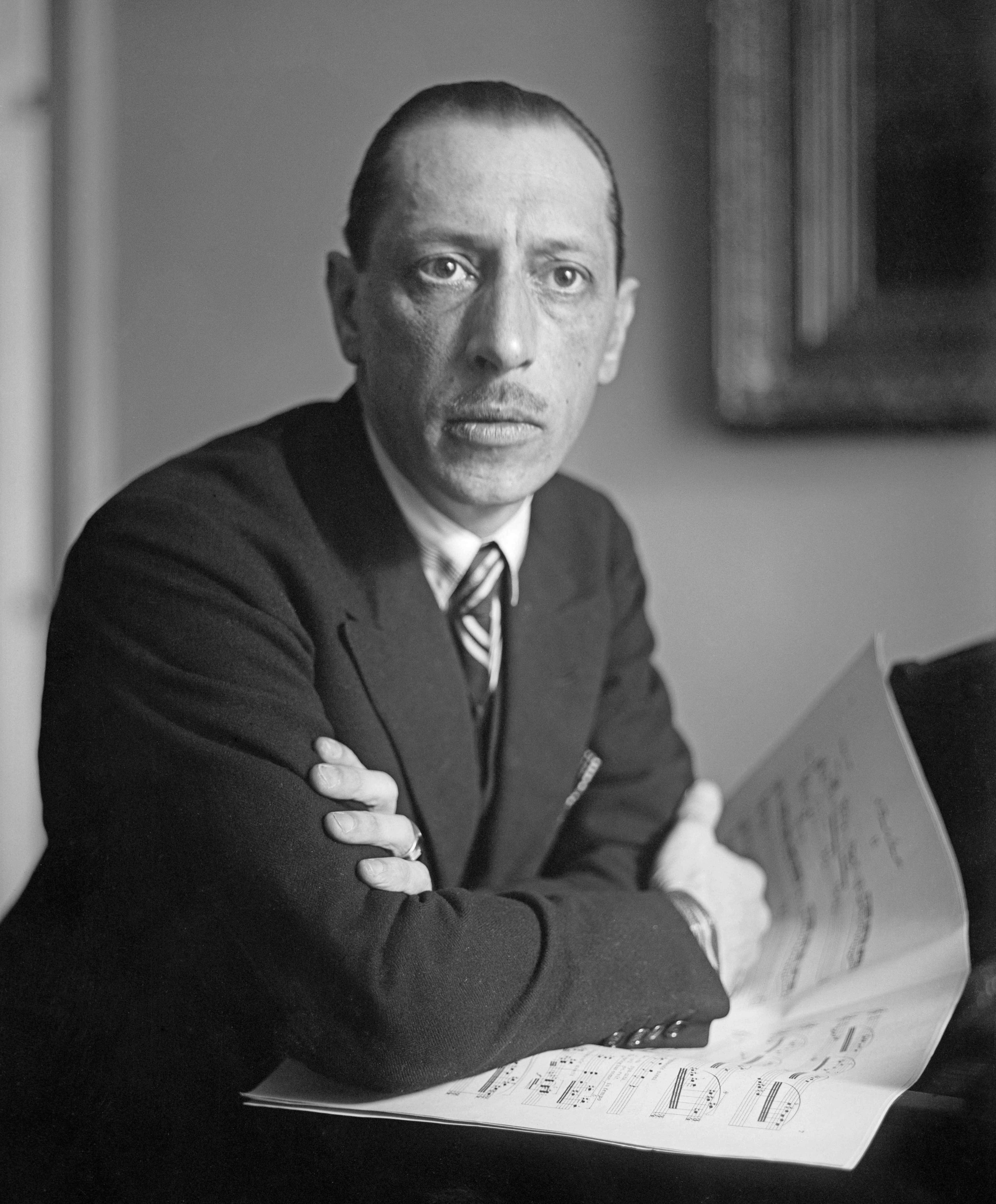Igor Stravinsky as a young man in round glasses and a dark suit, seated with his hands crossed, looking into the camera